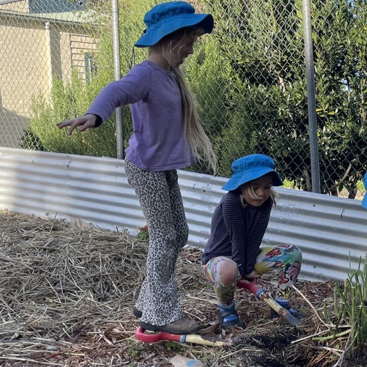 two children in blue hats stand and sit on a shovel handle to get something out if the garden together